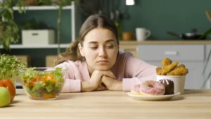 A woman in a pink top sits at a kitchen table, looking longingly at a plate of doughnuts and a cup containing fried snacks, with a bowl of salad nearby,