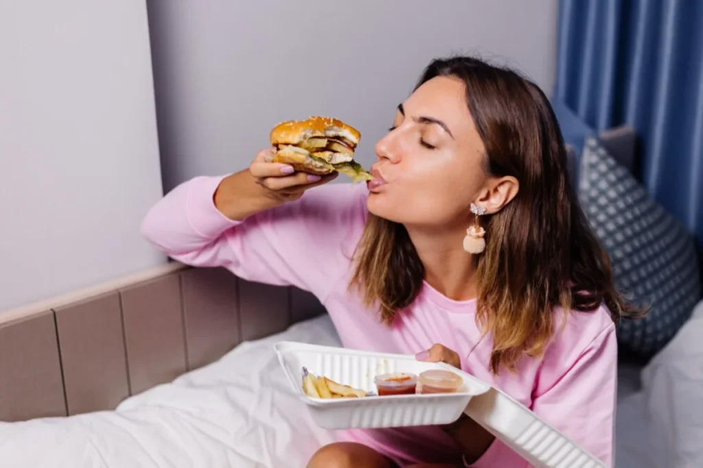 A woman in a pink sweatshirt sitting on a bed, eating a burger passionately while holding a takeaway tray with fries and sauces.