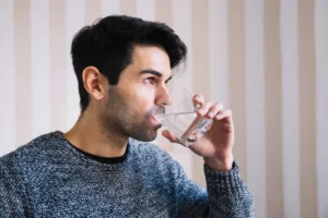 A man in a grey jumper drinking water from a clear glass