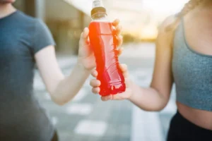 Two individuals in athletic wear sharing a red sports drink bottle