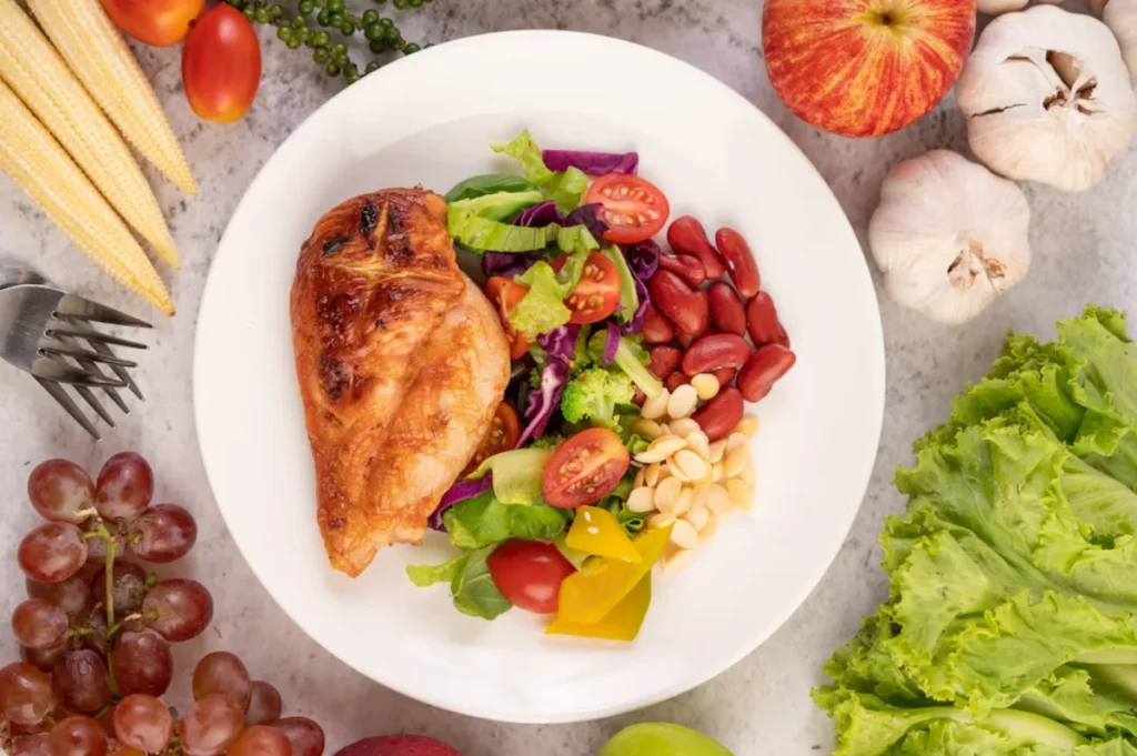 A balanced meal of grilled chicken breast served with a fresh vegetable salad containing cherry tomatoes, yellow bell pepper, broccoli, red cabbage, and pine nuts