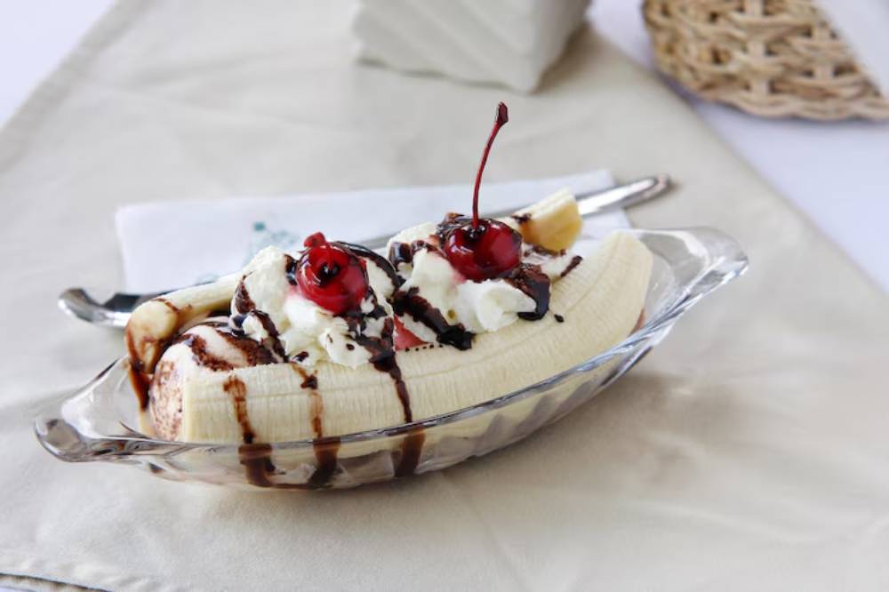 A banana split topped with whipped cream, chocolate syrup, and two maraschino cherries in a glass dish on a cream-colored tablecloth.