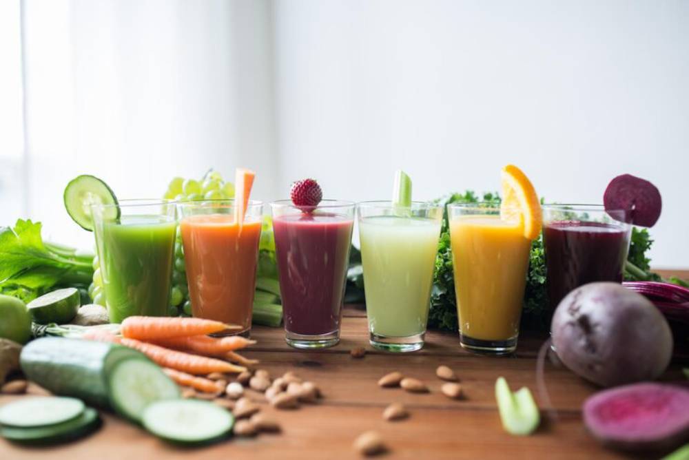 A vibrant display of six colorful juice glasses, surrounded by fresh fruits and vegetables on a wooden table.