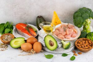Fresh ingredients for a healthy meal: salmon, chicken, avocado, broccoli, cheese, spinach, eggs, nuts, and olive oil on a textured background.