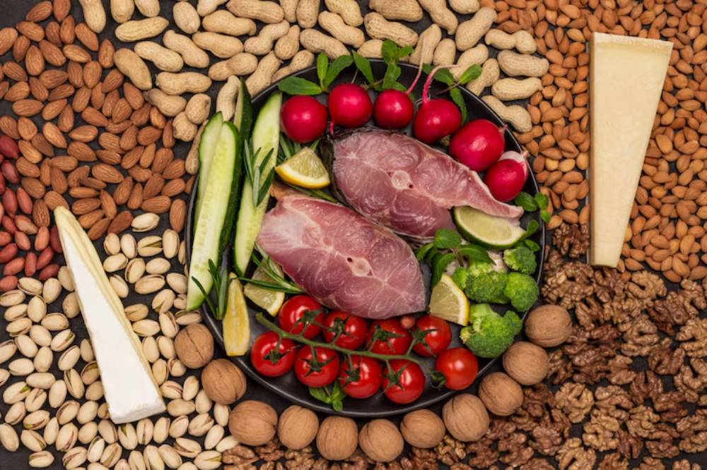 A plate of sliced fish surrounded by fresh vegetables, lemon, and assorted nuts, with wedges of cheese on the side.
