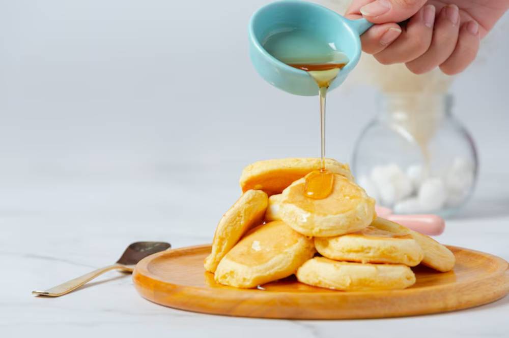 A hand pours syrup from a blue cup onto a stack of fluffy pancakes on a wooden plate, with a soft background.