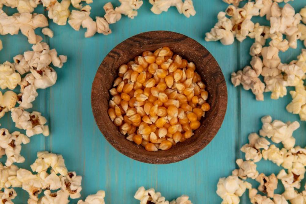 A wooden bowl filled with yellow popcorn kernels surrounded by scattered popped popcorn on a teal background.
