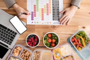 A person writes on a chart while surrounded by healthy meal prep, including fruits, vegetables, and a laptop. A smartphone is nearby.