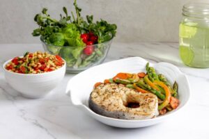A plate of grilled fish with sautéed vegetables, next to a bowl of tomato salad topped with seeds and a side of fresh greens.