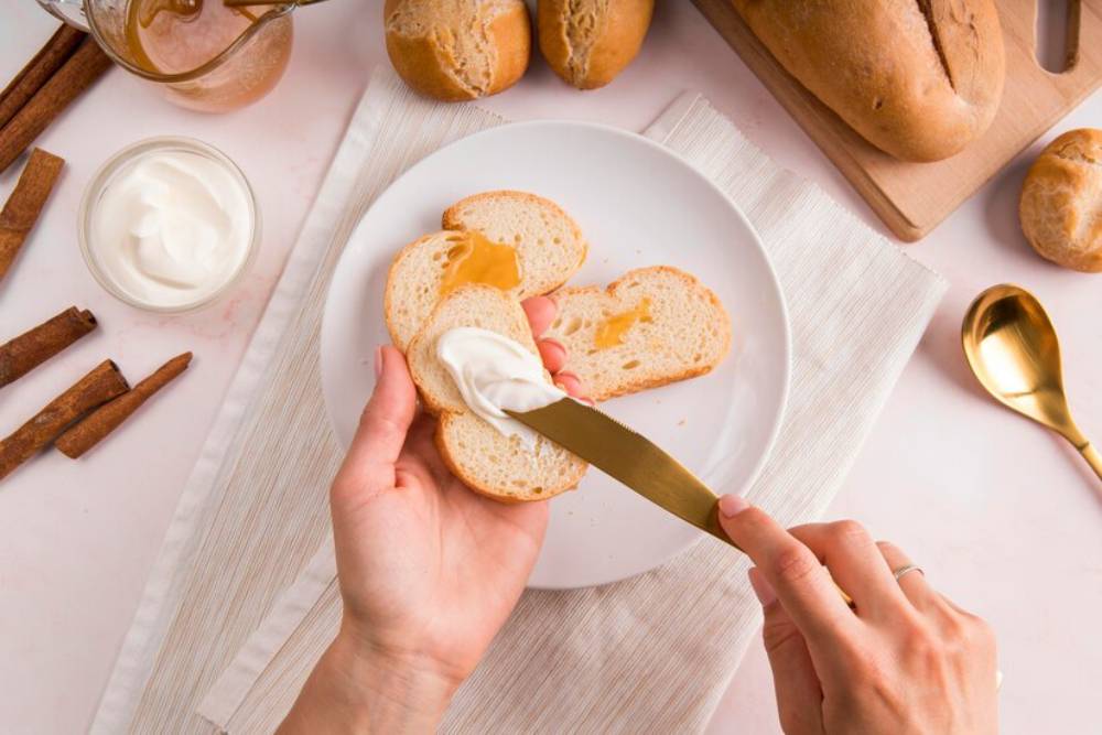 A hand spreads yogurt on a slice of bread topped with honey and a jar of yogurt placed on table.