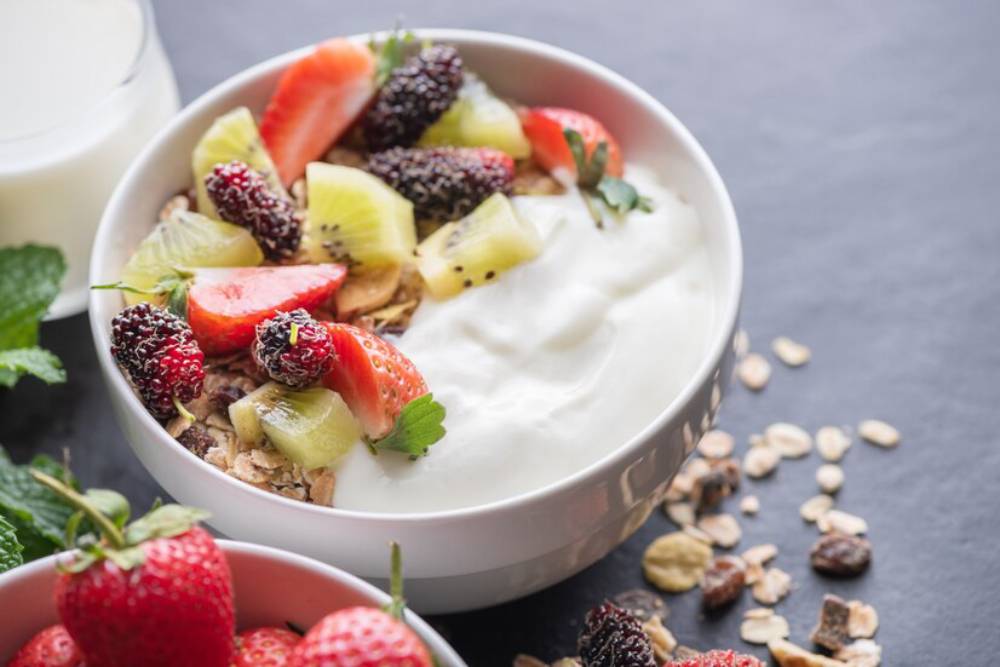 A bowl of yogurt topped with strawberries, kiwi, and blackberries, surrounded by granola and fresh strawberries on a dark surface.