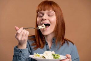 A woman with long red hair holds a fork in one hand and a plate of salad in the other, eating salad slowly.