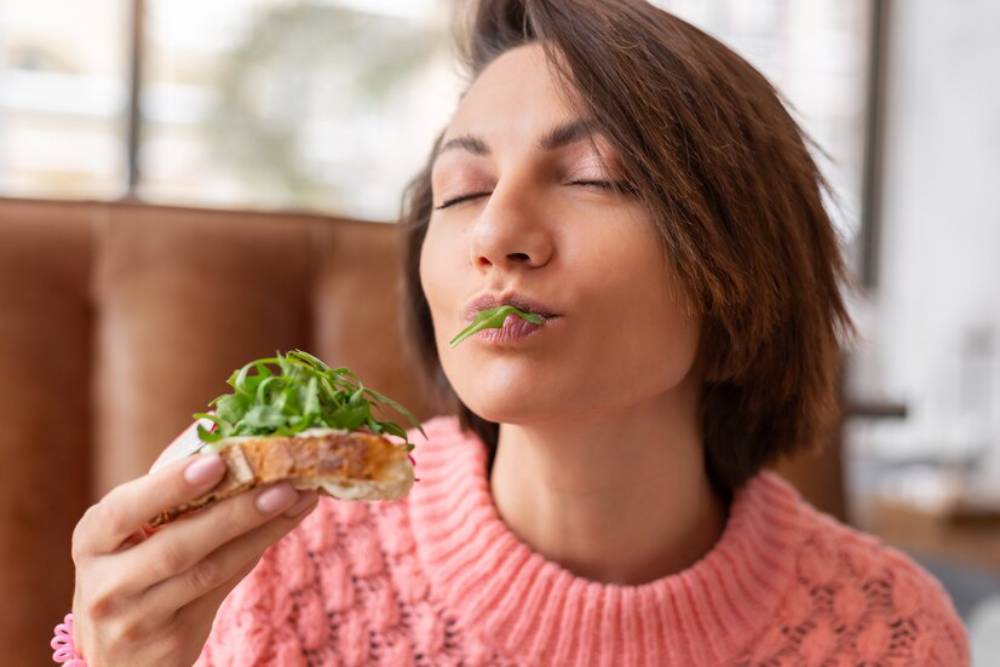 A person enjoys a slice of bread topped with fresh green arugula, wearing a cozy pink sweater in a café setting.