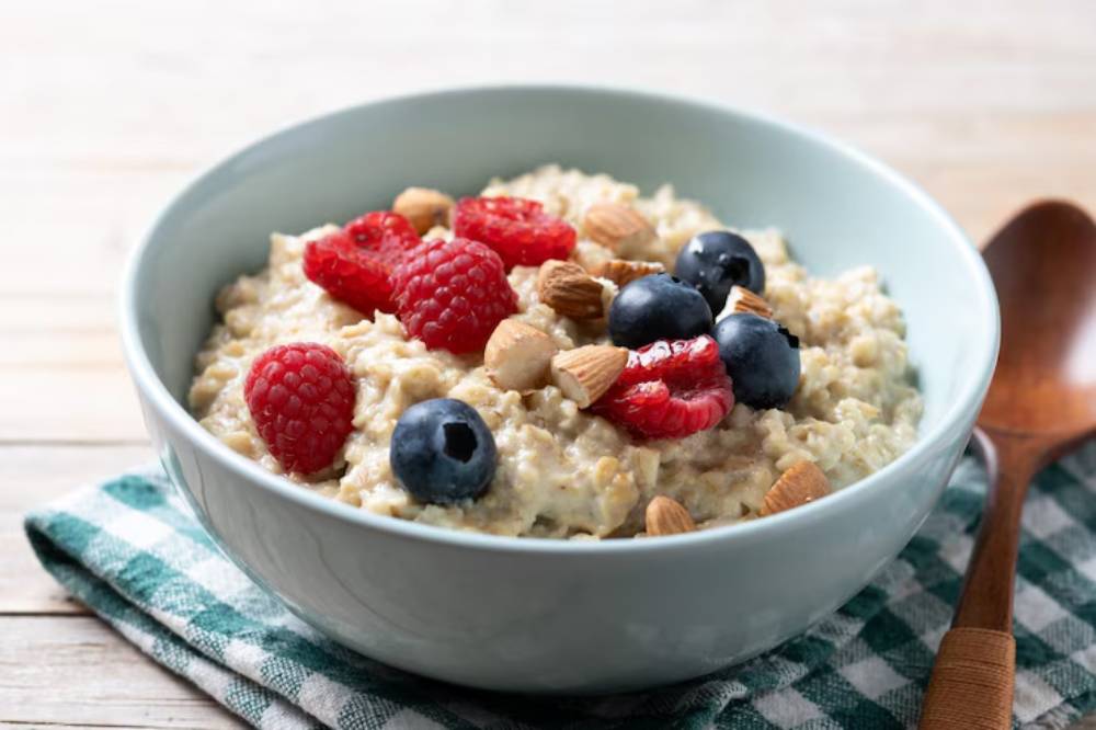 A bowl of oatmeal topped with fresh raspberries, blueberries, and almonds, resting on a checkered cloth with a wooden spoon nearby.