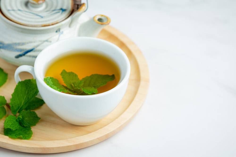 A steaming cup of green tea with fresh mint leaves, beside a ceramic teapot, on a wooden tray.