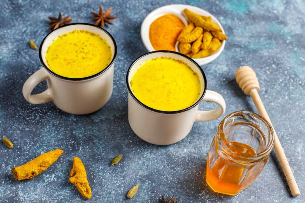Two mugs of turmeric latte on a textured blue surface, surrounded by spices, honey, and turmeric roots.