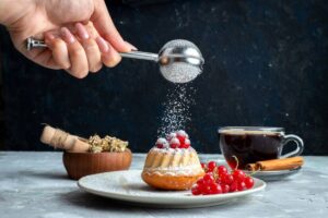 A hand sprinkles powdered sugar over a small bundt cake topped with red berries, alongside a cup of coffee and herbs on a plate.
