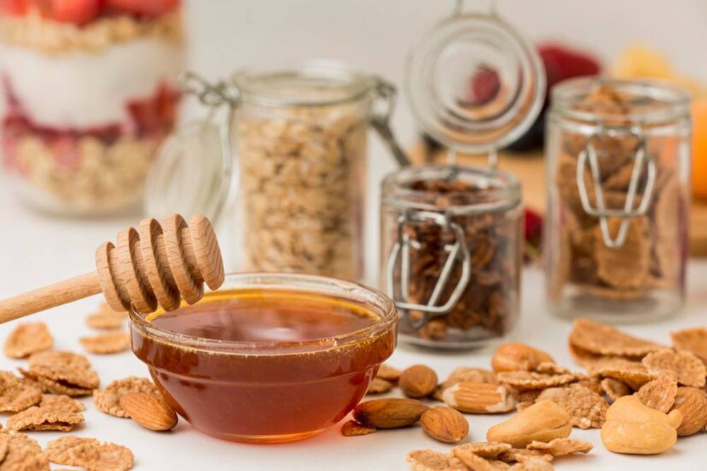 A bowl of honey with a wooden dipper, surrounded by nuts and cereal, with jars of oats and snacks in the background.