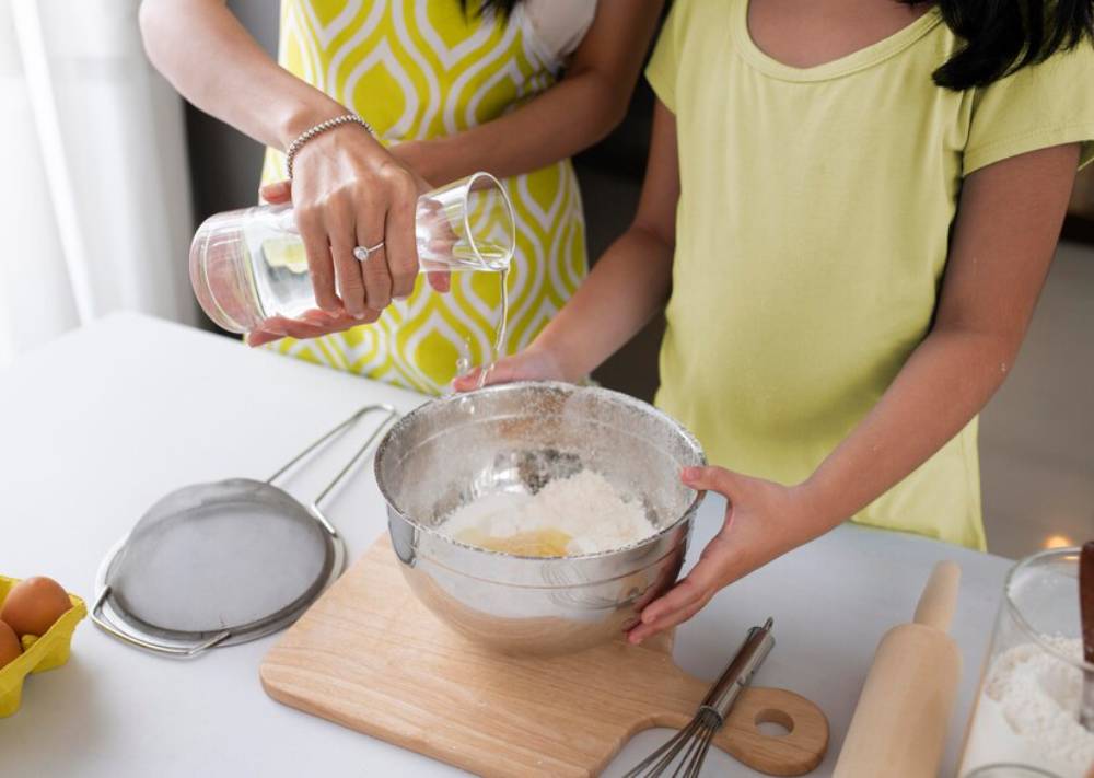 A mother and daughter are baking together, with the mother pouring water into a mixing bowl while the girl assists.