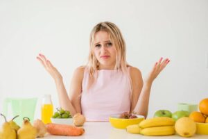 A person in a pink top gestures with hands and confused face, surrounded by an array of colorful fruits and vegetables on a white table.