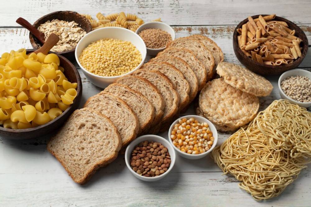 A variety of grains, legumes, and pasta displayed on a wooden surface, including bread, rice cakes, and assorted seeds in bowls.