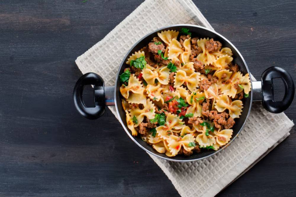 A skillet filled with farfalle pasta mixed with ground meat and garnished with fresh herbs, resting on a beige cloth.