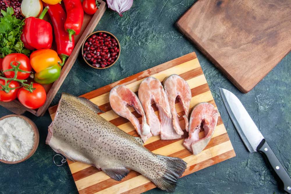 A wooden cutting board with fish fillets, a whole fish, and a variety of fresh vegetables including peppers, tomatoes, and herbs.