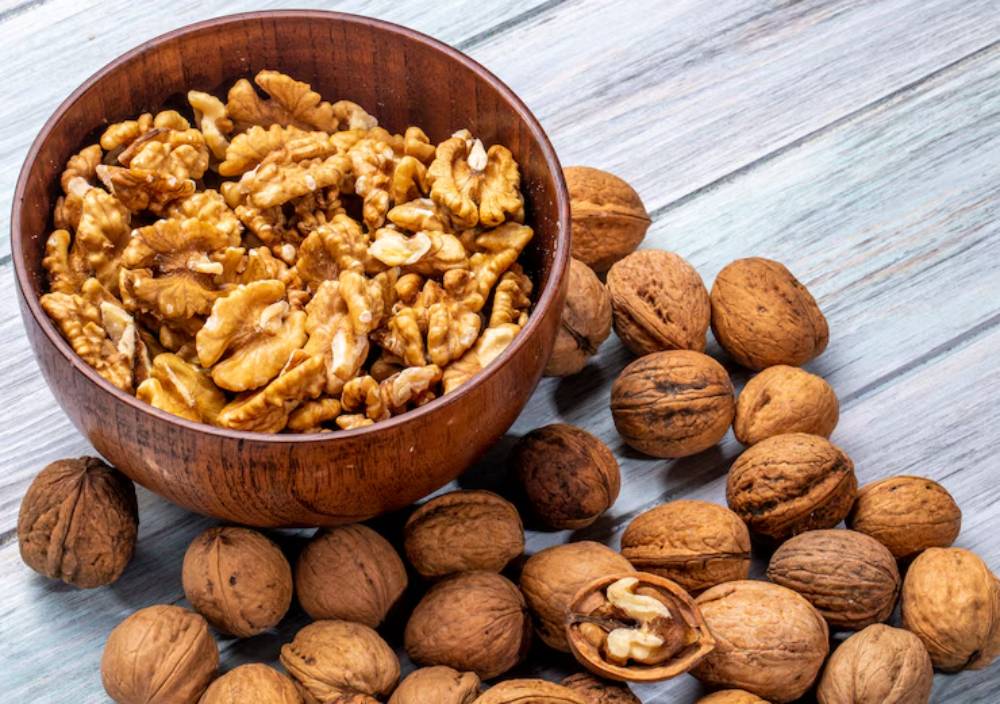 A wooden bowl filled with shelled walnuts rests among whole walnuts on a light wooden surface.