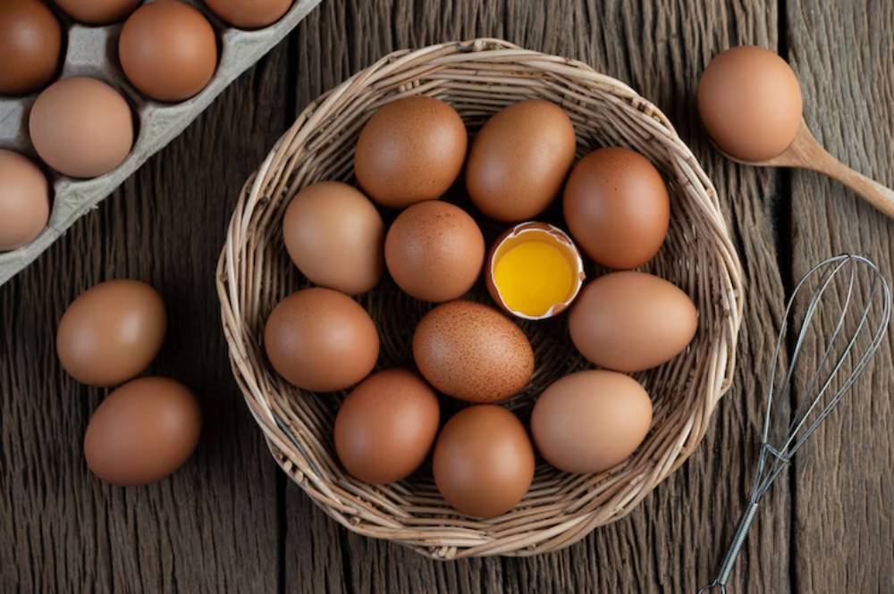 A woven basket filled with brown eggs, one cracked open revealing a yellow yolk, accompanied by a whisk and a wooden spoon.
