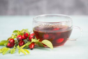A clear glass cup filled with steaming rosehip tea next to vibrant red rosehips and green leaves on a light blue surface.